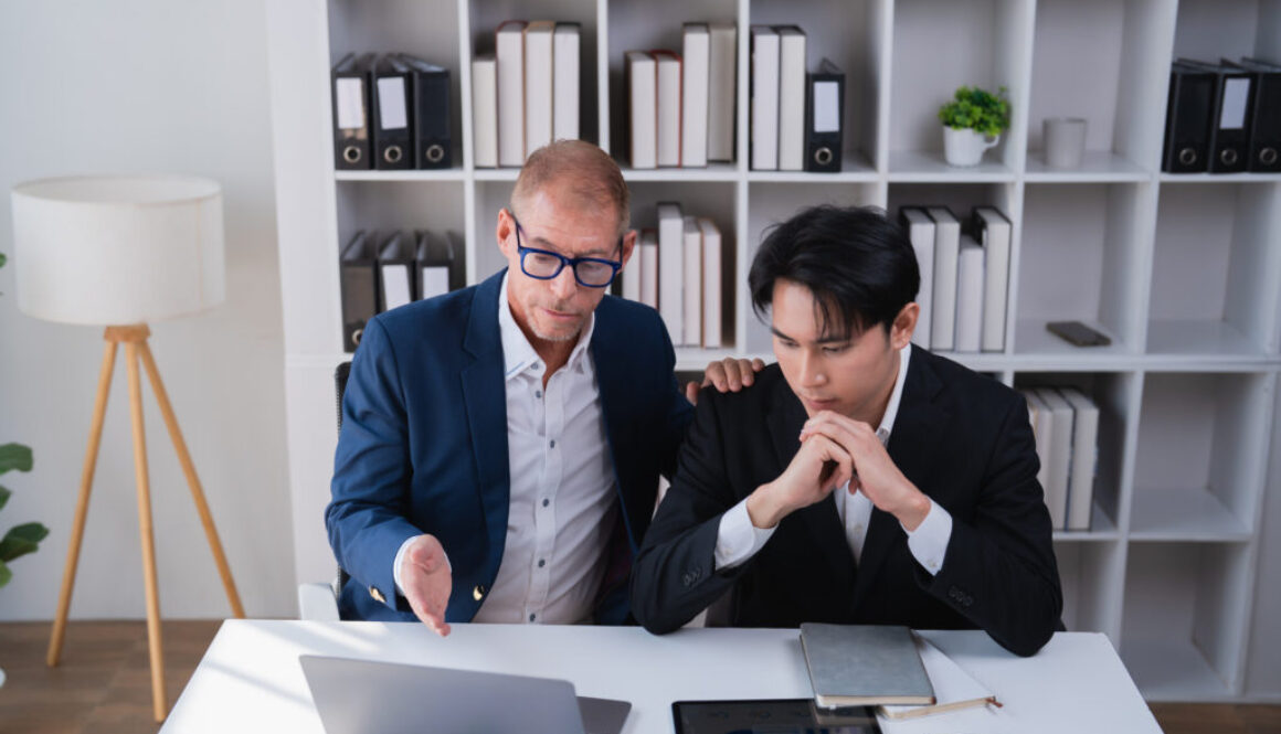 Two distinct generation businessmen collaborating and discussing financial data on a laptop, providing guidance and support in a modern office workplace setting